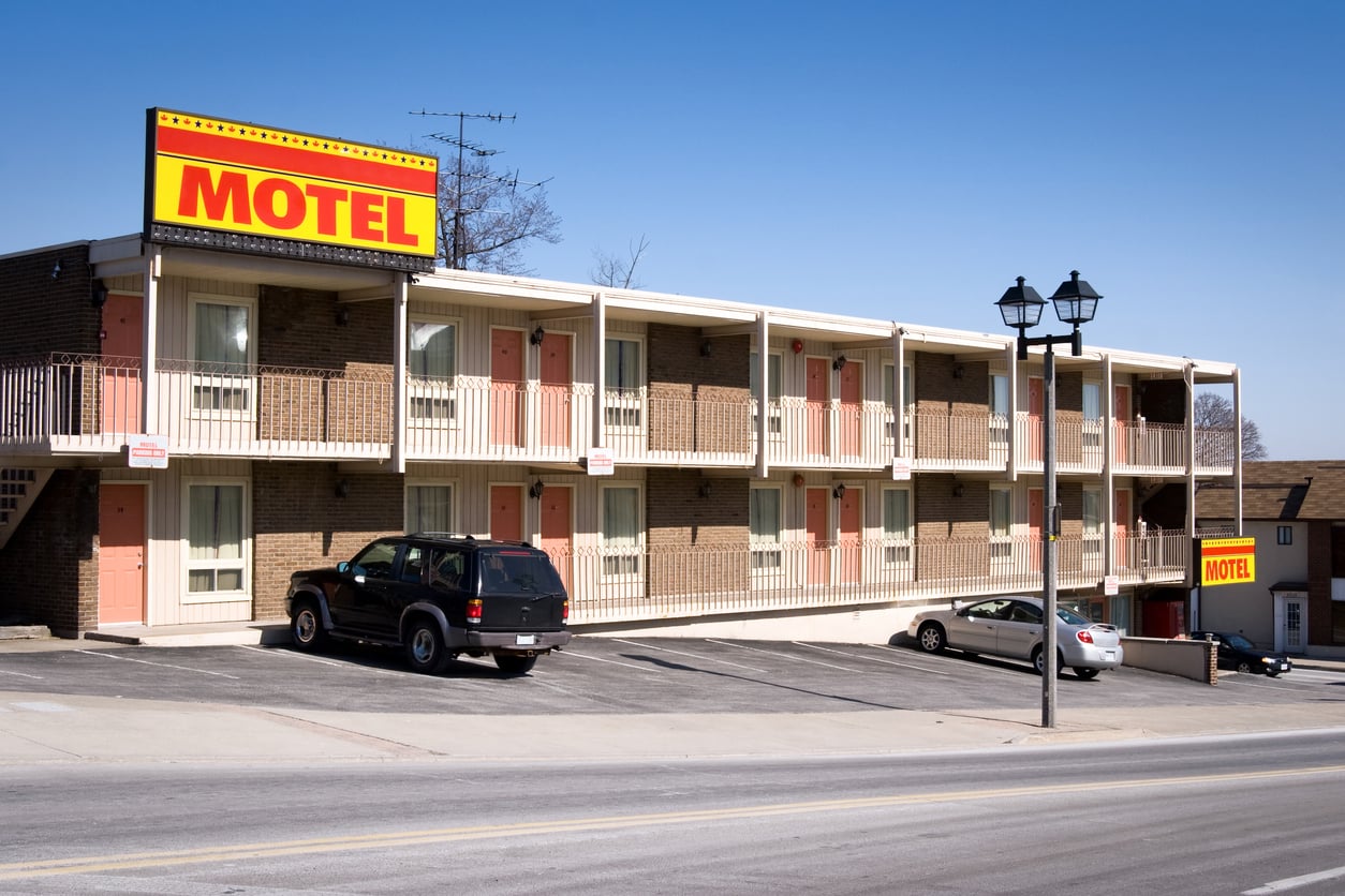 A two-story brick motel with a yellow and red “MOTEL” sign, outdoor balconies, and parked cars in front on a sunny day—an inviting spot for a relaxing vacation.