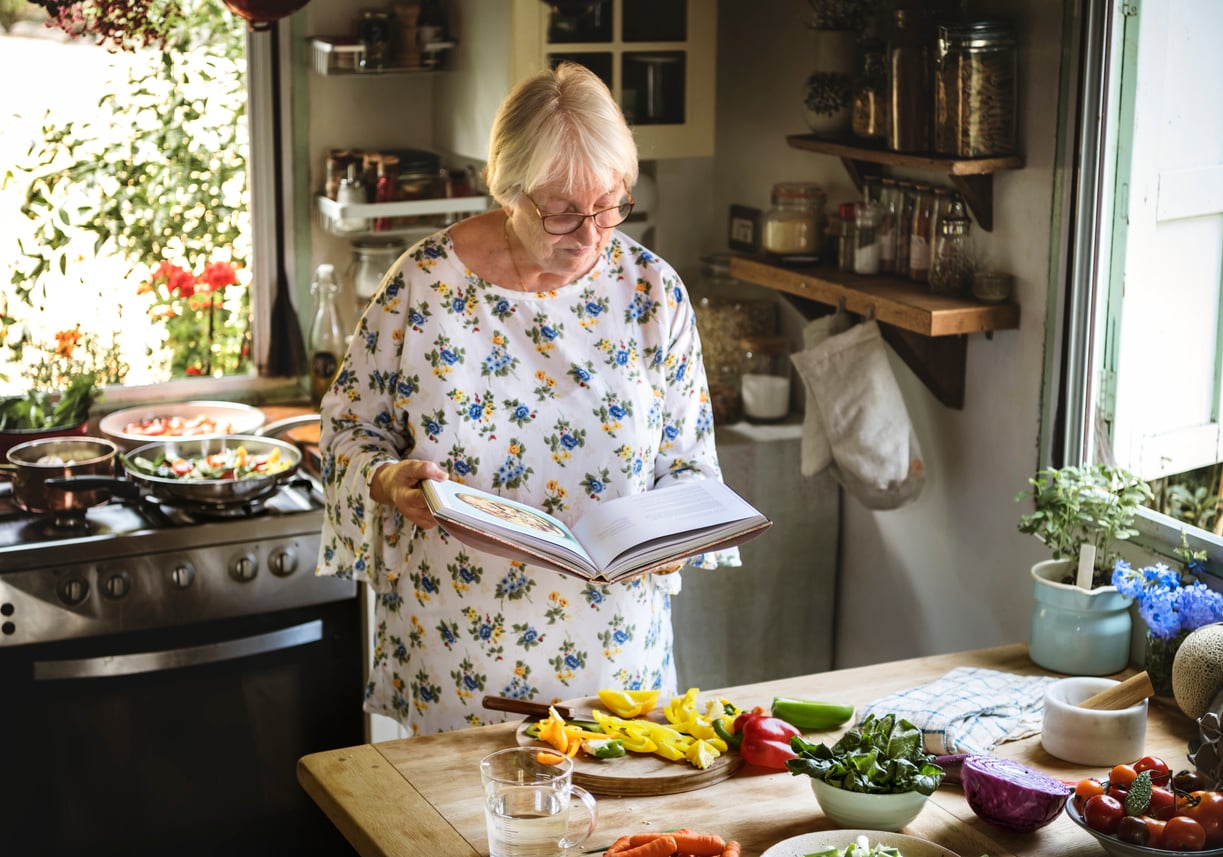 An older woman in a floral shirt reads a cookbook in a bright kitchen, surrounded by fresh vegetables and cooking utensils on the counter.