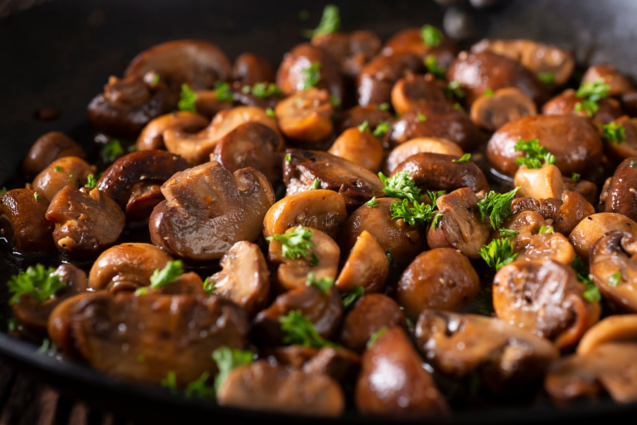 Sautéed mushrooms, often considered one of those foods people learn to appreciate later in life, are garnished with chopped parsley in a black skillet. The mushrooms are golden brown and glistening, with fresh green parsley sprinkled on top.