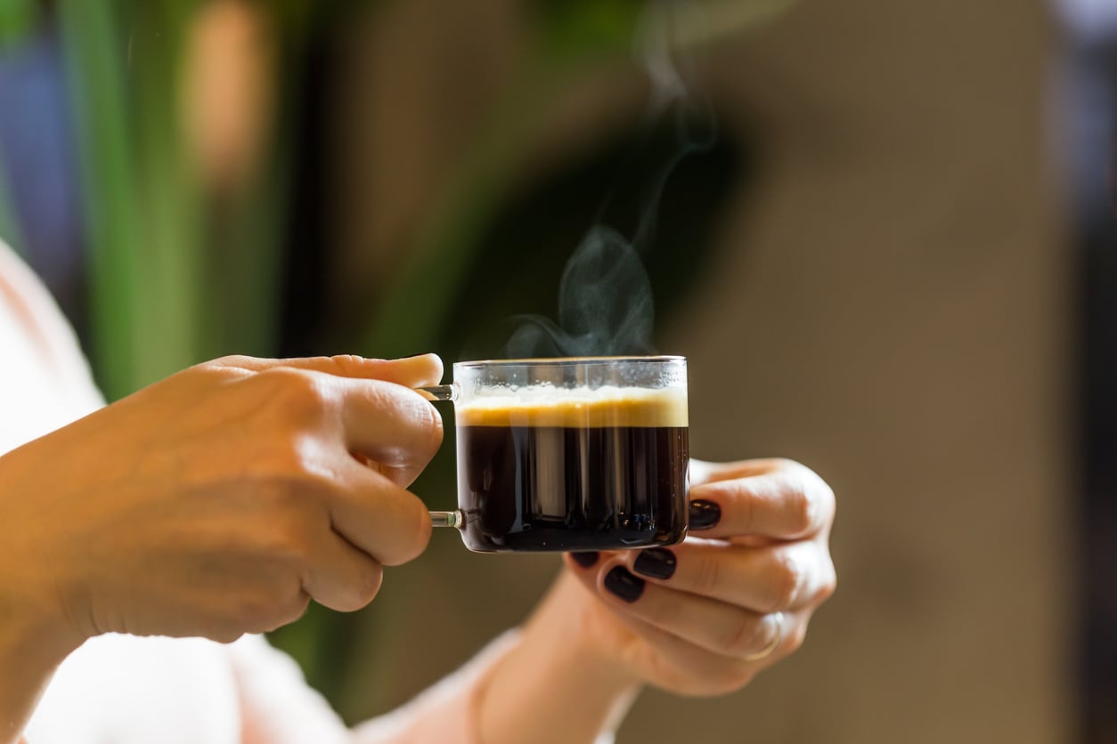 A person holding a small glass cup of hot espresso, one of those foods people learn to appreciate later in life, with steam rising from the drink, against a blurred background with green and brown tones.