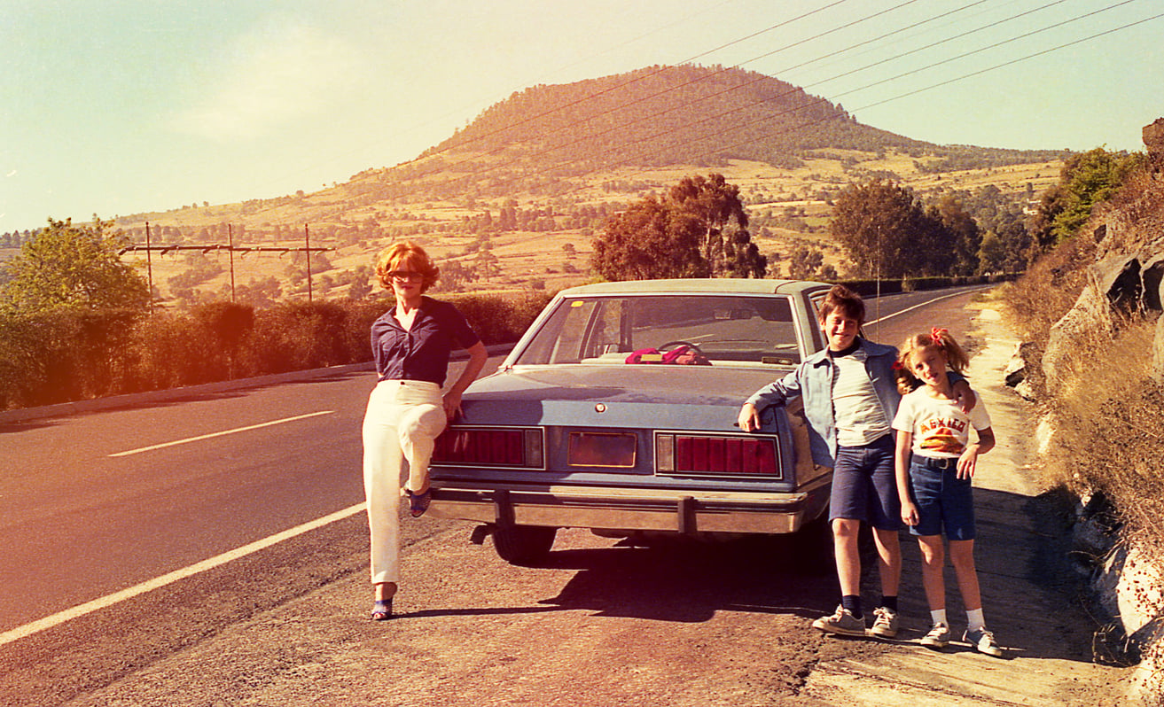 A woman and two children stand by a parked car on a rural road with a hill and trees in the background on a sunny day, capturing the essence of travel in the 60s and 70s.