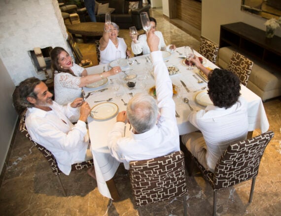 Five people in white clothing sit around a dining table, raising glasses in a toast and smiling. The table is set formally with plates, glasses, and a few dishes, in a modern room with patterned chairs and marble floor.