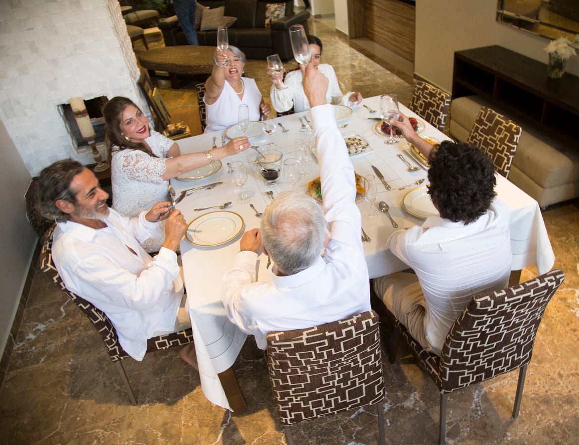 Five people in white clothing sit around a dining table, raising glasses in a toast and smiling. The table is set formally with plates, glasses, and a few dishes, in a modern room with patterned chairs and marble floor.