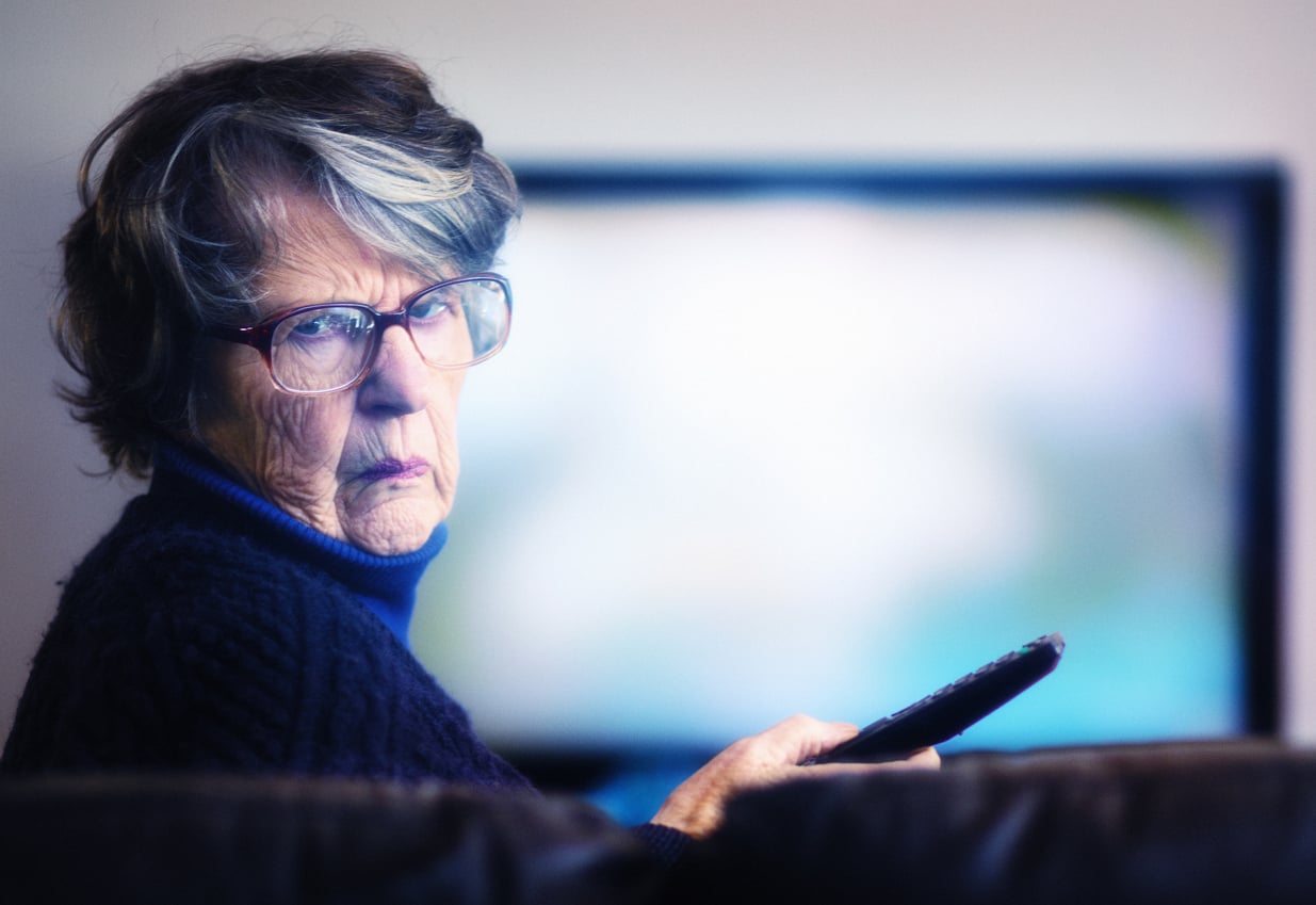 An elderly woman with glasses and gray hair holds a remote control and looks seriously at the camera, sitting on a couch with a blurry television screen in the background.