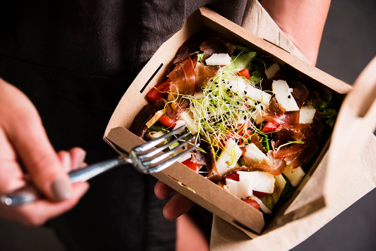 A person holds a takeout box with a fresh salad topped with sliced cured meat, shaved cheese, and sprouts, while using a fork to pick up a bite.