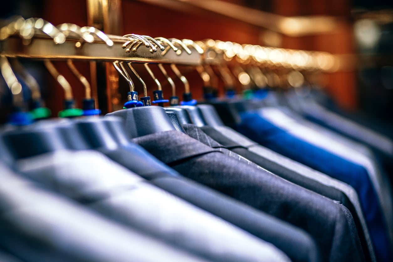 A row of suits in various shades hangs neatly on hangers, displayed on a gold clothing rack in a store or closet.