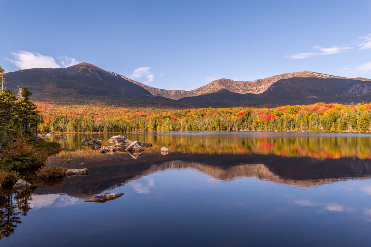A calm lake reflects autumn trees and distant mountains under a clear blue sky. Rocks and evergreens line the shore, and colorful fall foliage covers the forest.