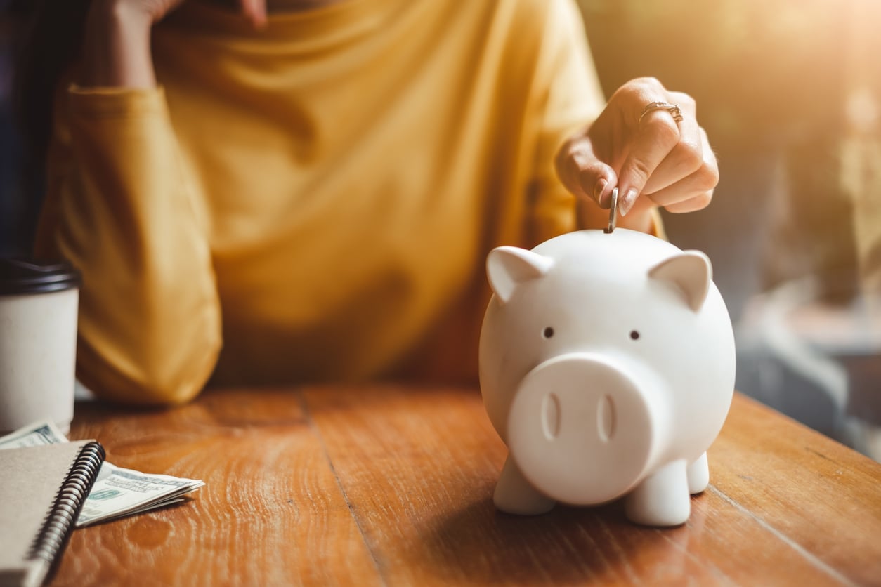 A person in a yellow sweater puts a coin into a white piggy bank on a wooden table, demonstrating positive retirement money habits, with a coffee cup, notebook, and cash nearby.