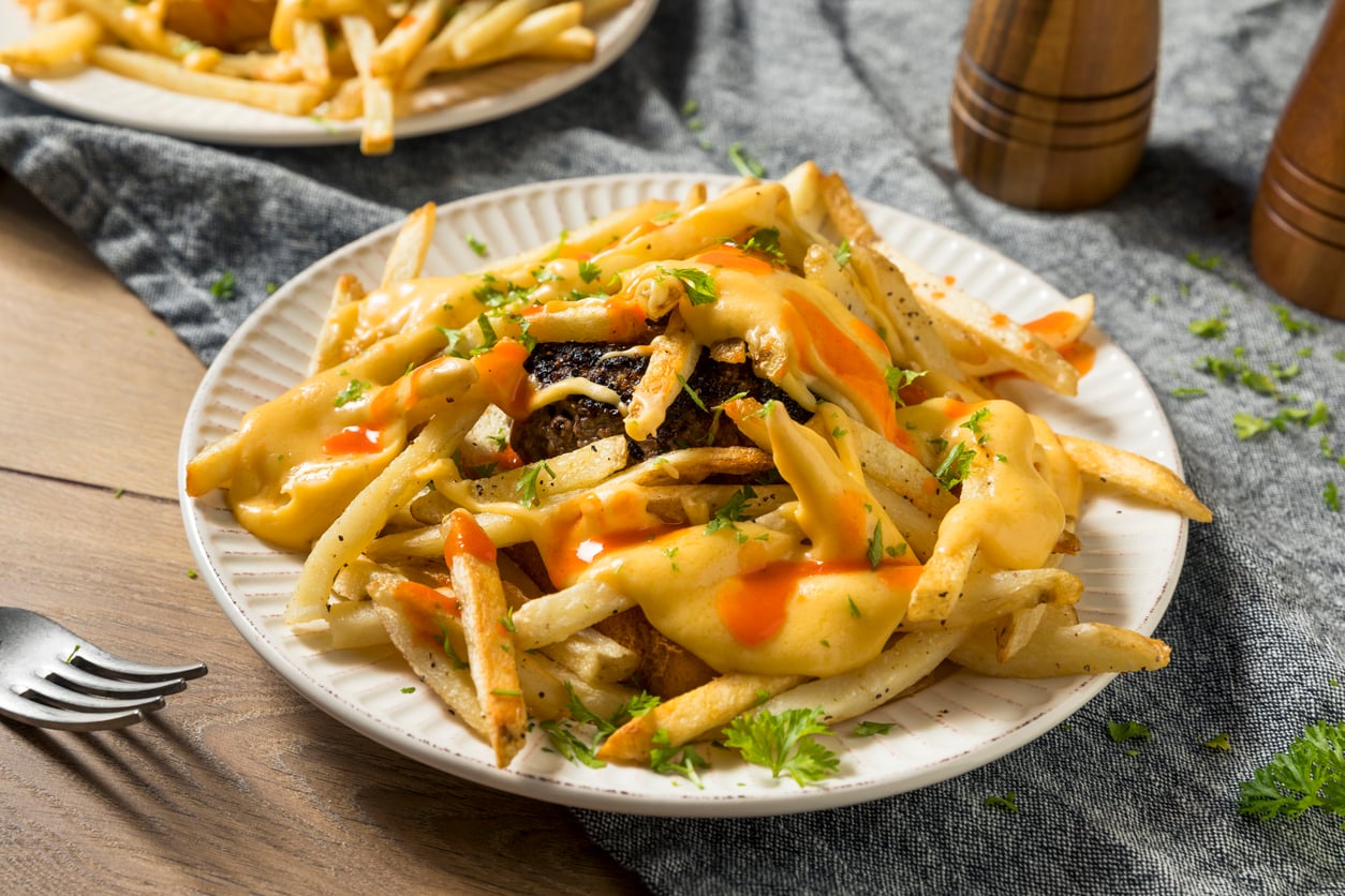 A plate of French fries topped with melted cheese sauce, hot sauce, and chopped herbs—one of the restaurant specialties—served on a white plate with a fork nearby on a wooden table.