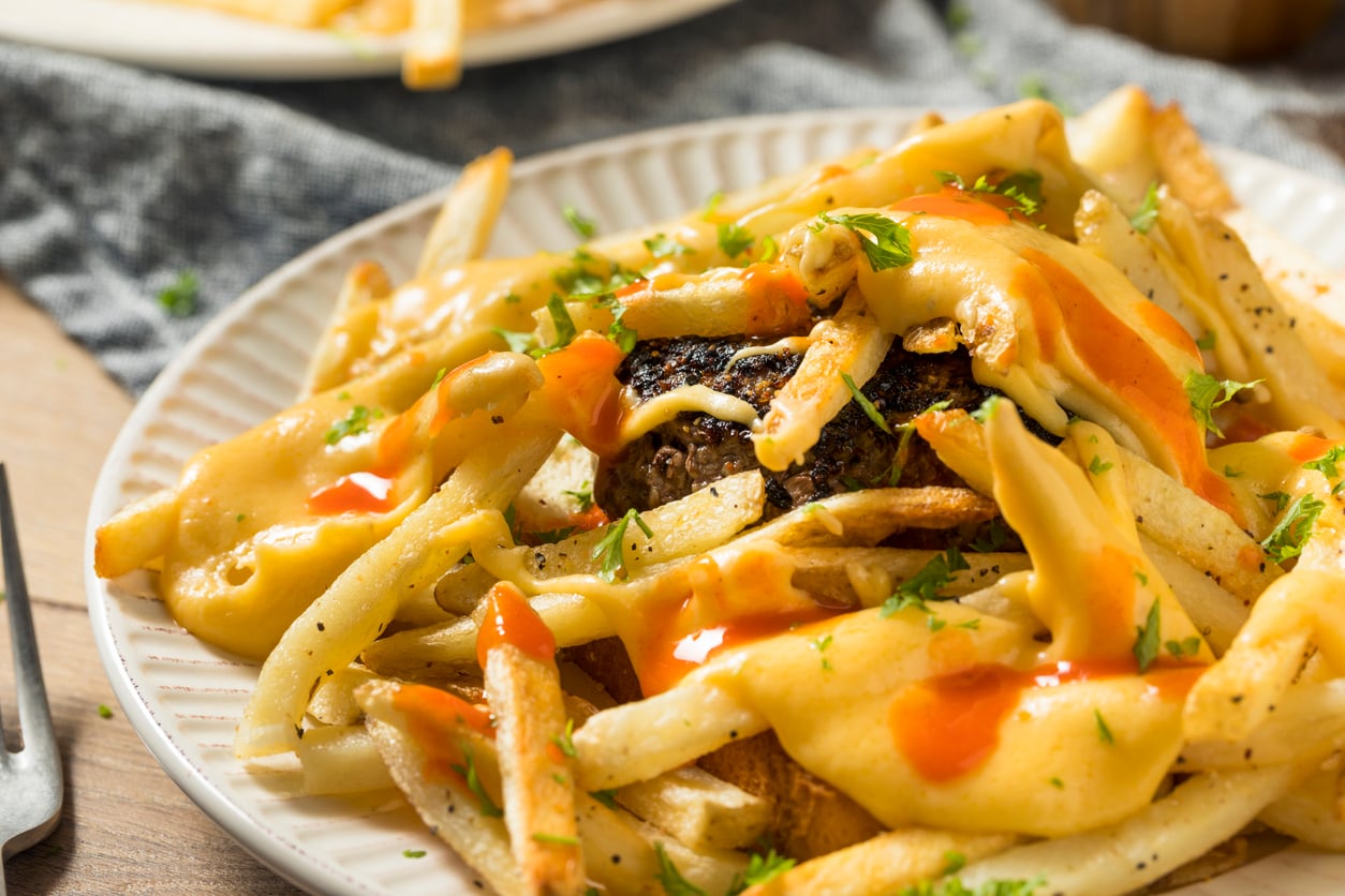 A plate of fries topped with melted cheese, gravy, hot sauce, chopped herbs, and a hamburger patty, served on a white dish.
