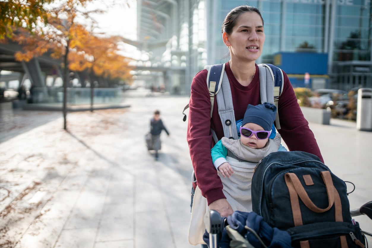 A woman pushes a stroller outdoors with a baby in a carrier wearing sunglasses and a hat. A child follows behind pulling a suitcase, and autumn trees line the sidewalk near a modern building.
