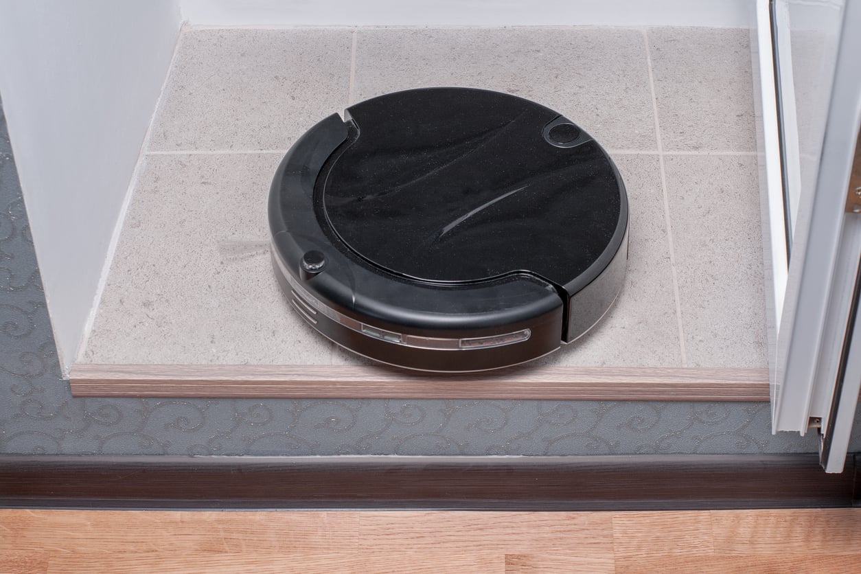 A black robotic vacuum cleaner sits on a tiled floor near a step, appearing to pause before reaching the edge.