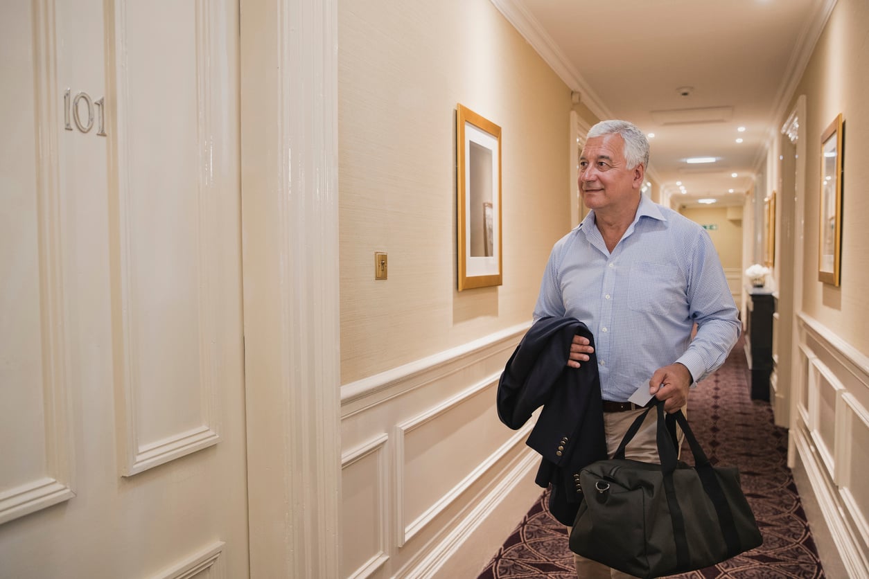 An older man in a light blue shirt carries a black bag and a jacket while walking down a hotel hallway, approaching room 101.