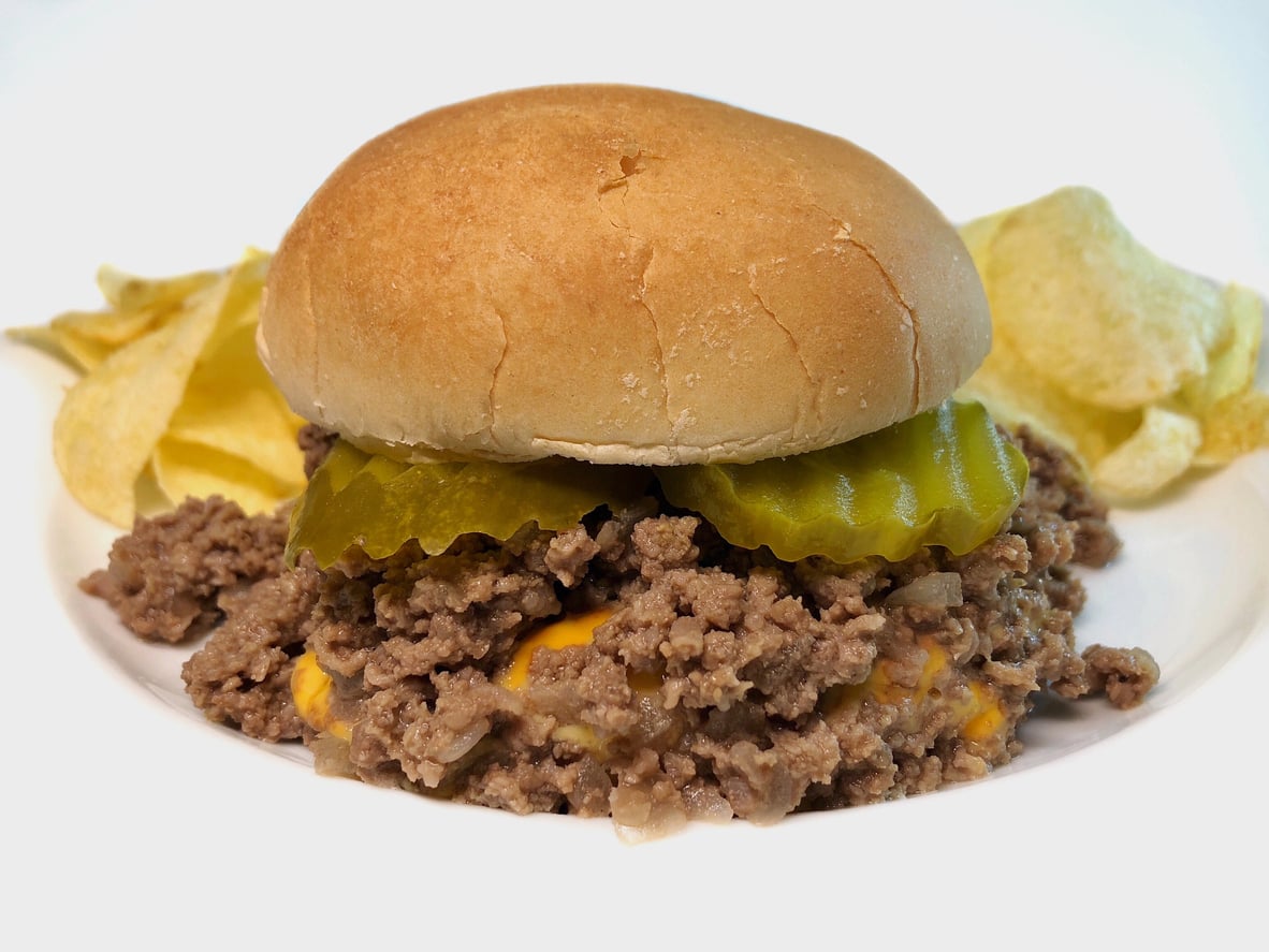 A sloppy joe sandwich with ground beef, melted cheese, and pickles on a bun, served with potato chips on a white plate.
