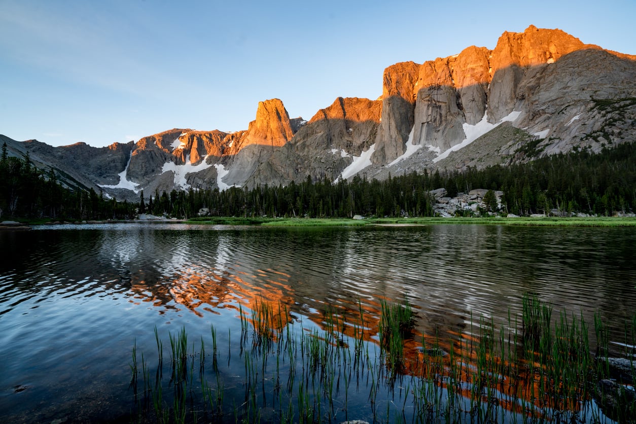 A mountain range with snow patches is illuminated by golden sunlight, reflecting in a calm lake with grasses in the foreground and pine trees along the shore.