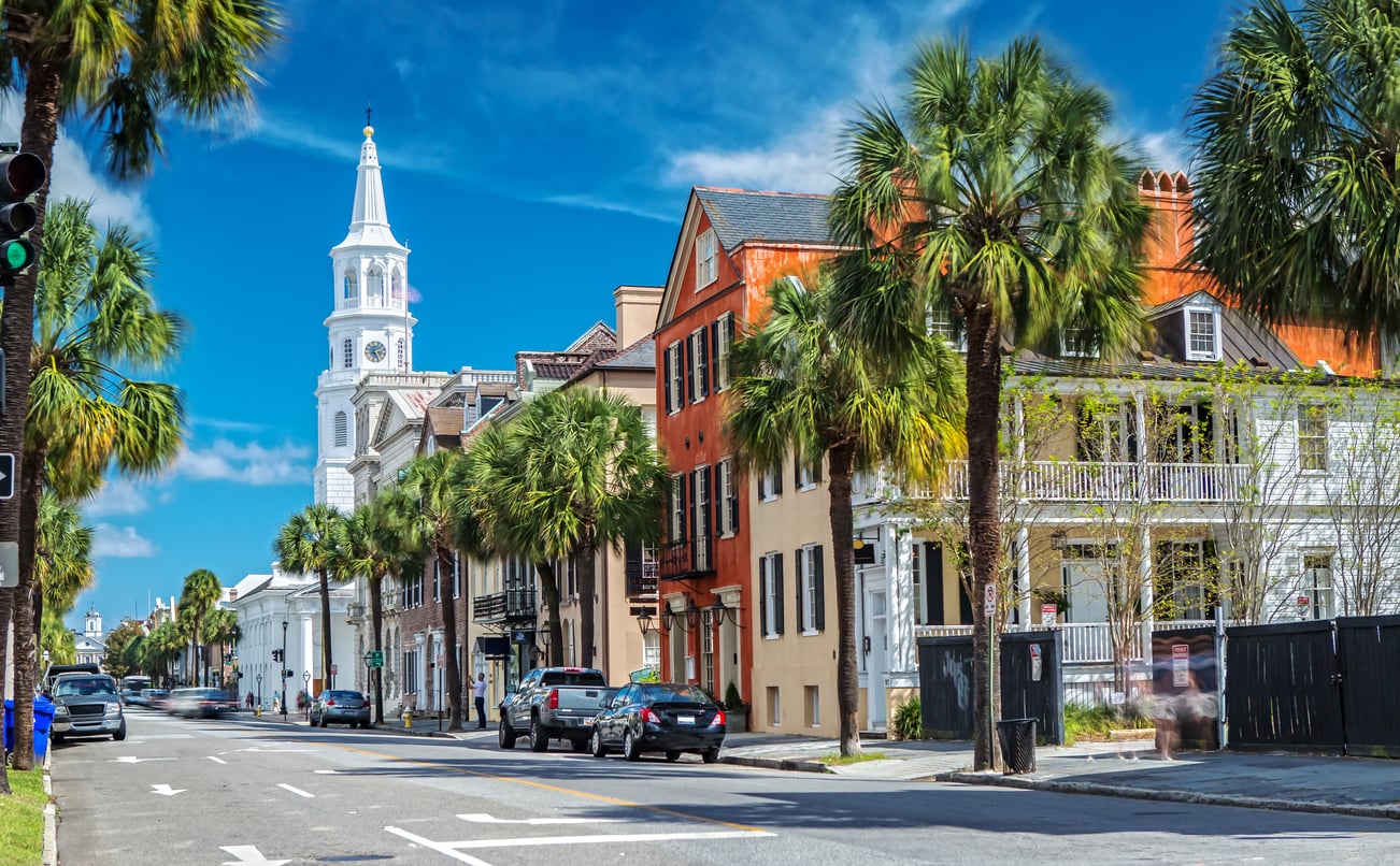 A street lined with palm trees, colorful historic buildings, parked cars, and a white church with a tall steeple under a blue sky.