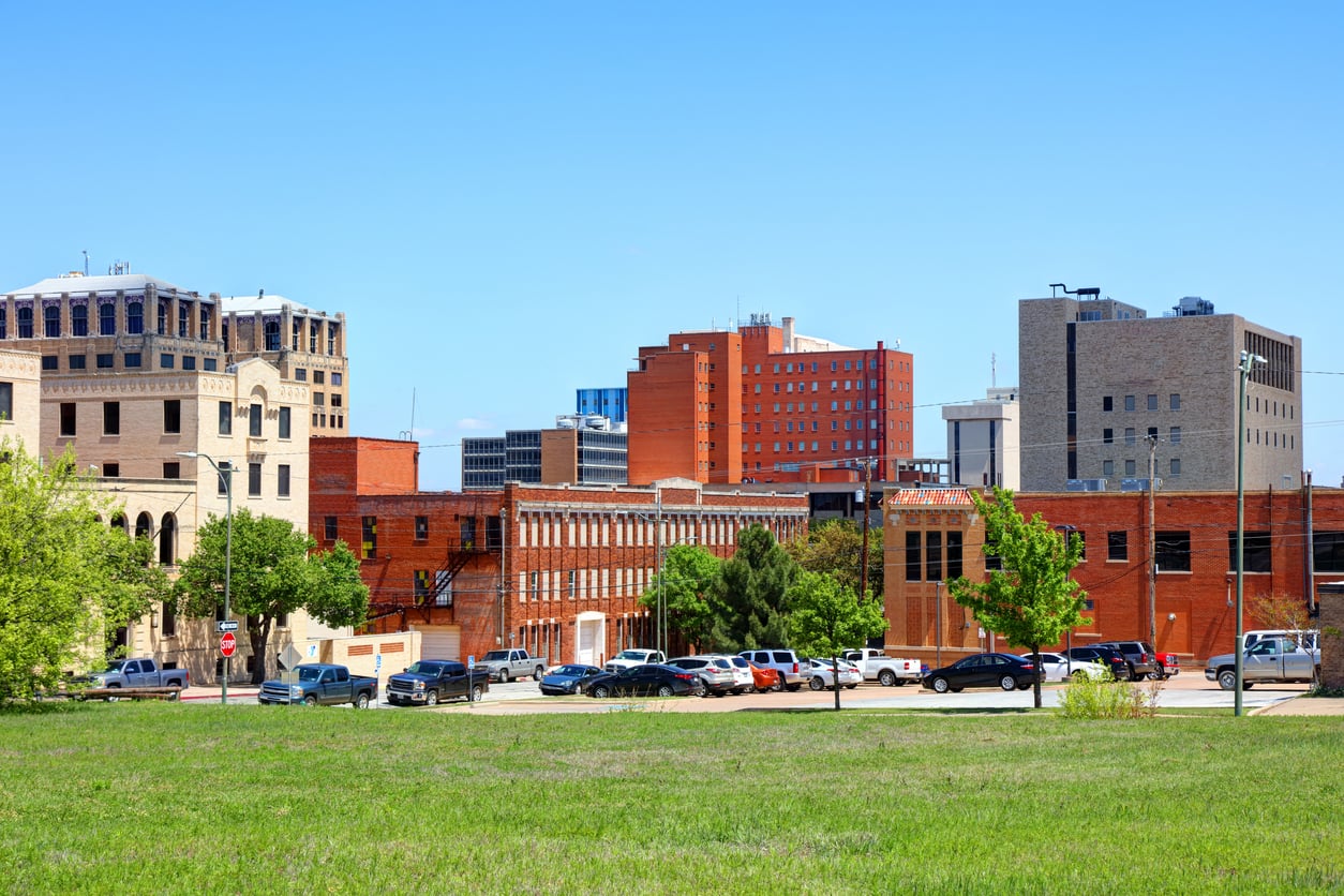 A grassy field in the foreground with parked cars and tall, red-brick buildings under a clear blue sky in the background.