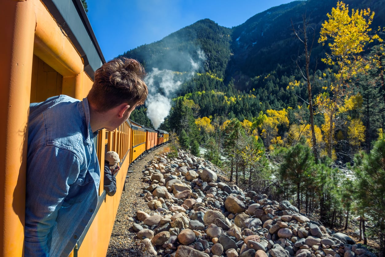 A person leans out of a yellow train, looking at the rocky landscape and green and yellow trees on a sunny day in the mountains. Steam rises from the train engine ahead.