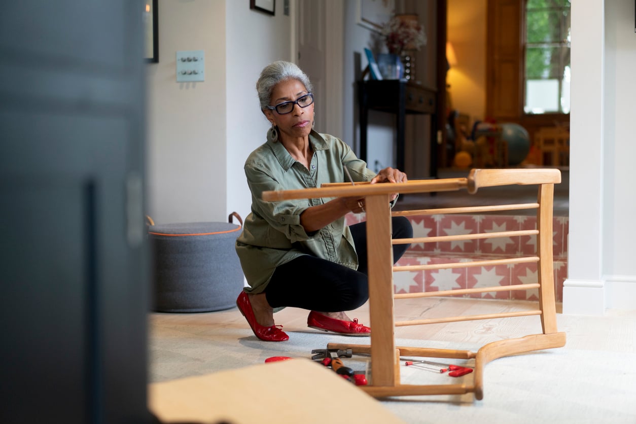 A woman with gray hair and glasses is kneeling on the floor, assembling a wooden table in a bright room. Tools are scattered nearby, and she appears focused on her task.