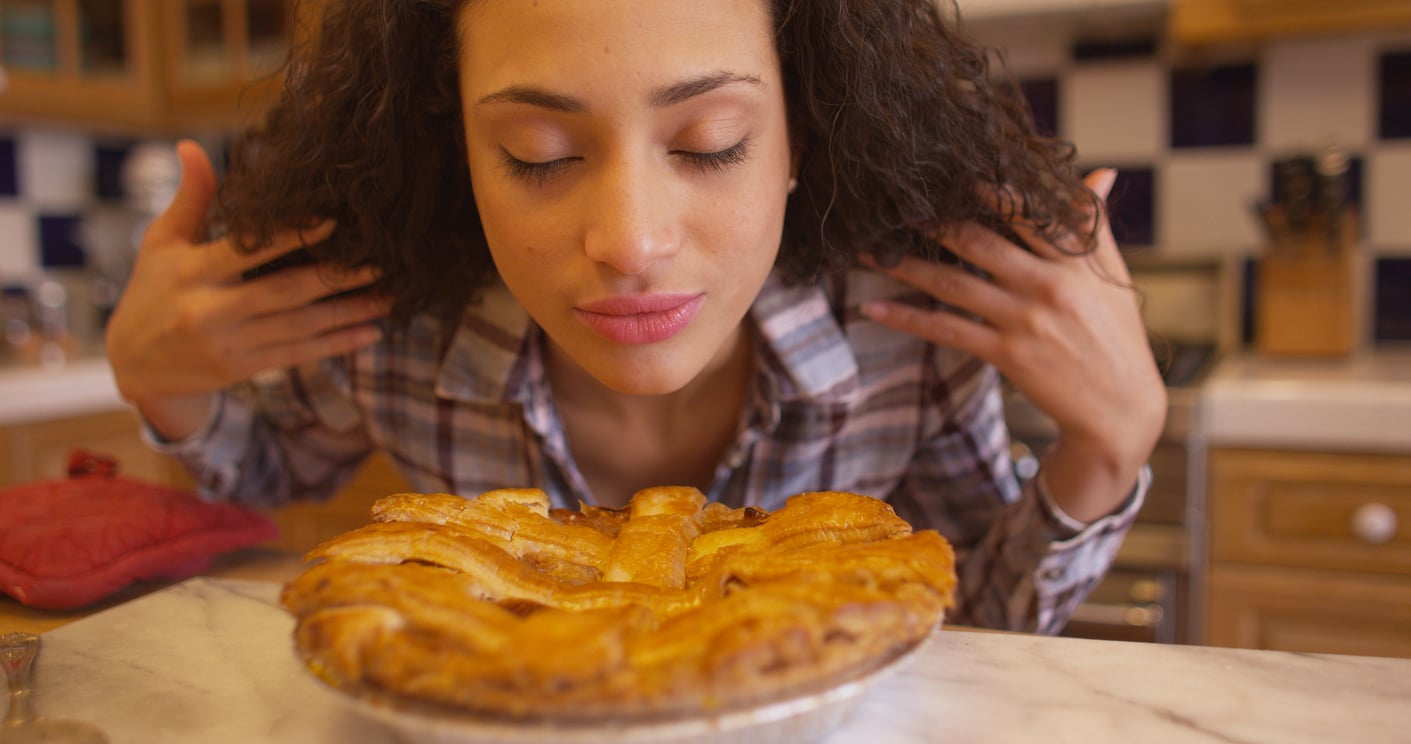 A woman with curly hair closes her eyes and inhales the aroma of a freshly baked pie, sitting on a kitchen counter in front of her.