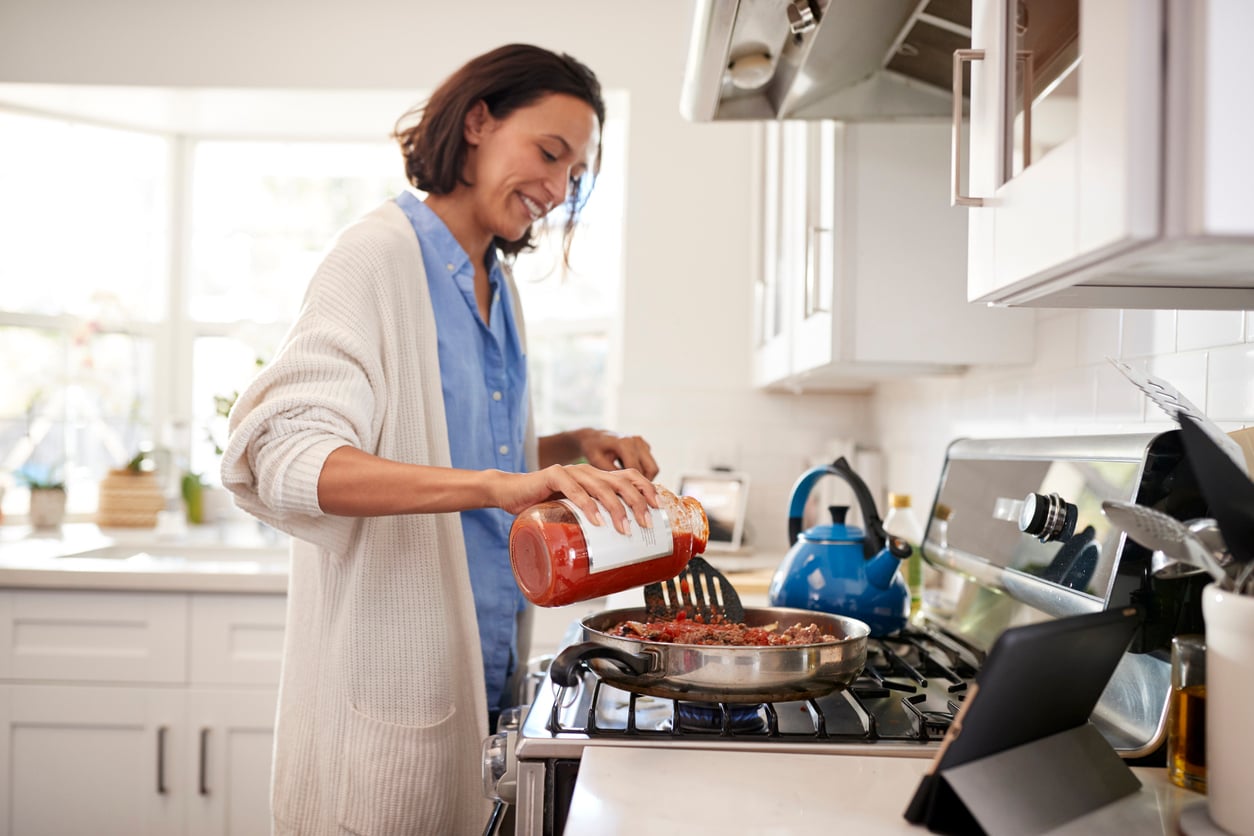 A person in a cozy kitchen pours tomato sauce from a jar into a skillet on the stove, smiling as they cook. A blue kettle and tablet are on the counter nearby.