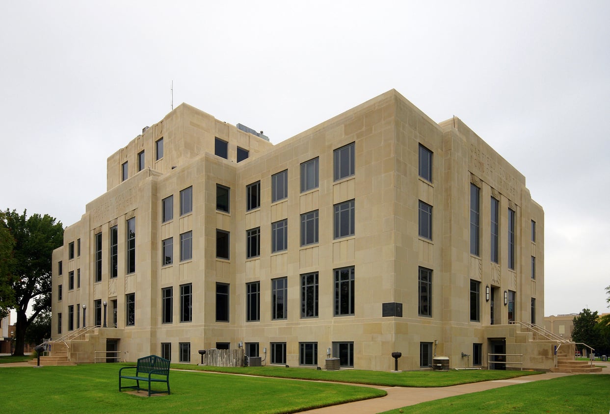 A large, beige stone courthouse with multiple windows stands on a manicured lawn with a bench and trees nearby under a cloudy sky.