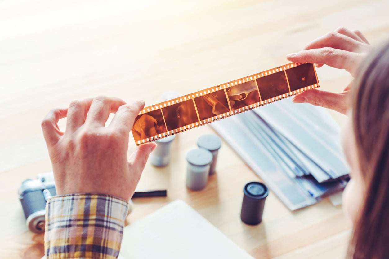 A person holds up a strip of photographic film negatives, examining it in the light. Film canisters, plastic sleeves, and a camera are on the wooden table below.