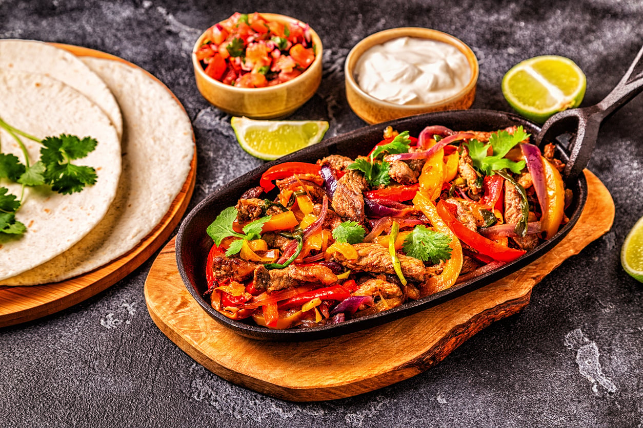 A skillet of chicken fajitas with bell peppers and onions, served with flour tortillas, salsa, sour cream, lime wedges, and cilantro on a wooden board.