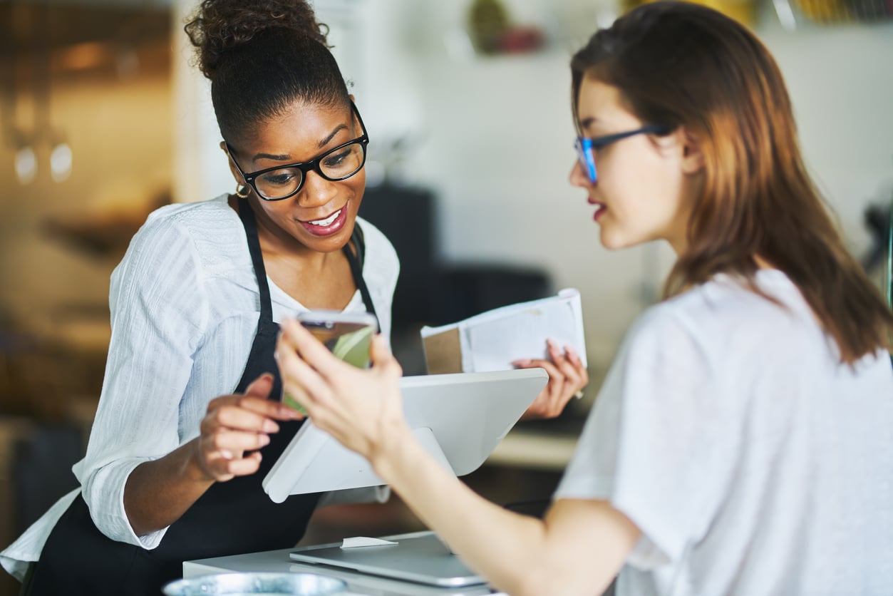 Two women interact at a counter; one wears an apron and smiles while the other holds a smartphone and shows the screen, both appearing engaged in a friendly conversation or transaction in a bright setting.