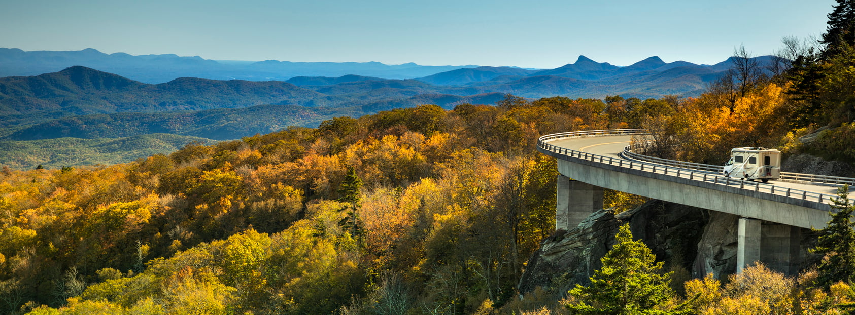 A white RV drives on a bridge curving over colorful autumn trees, with rolling blue mountains and a clear sky—perfect for trips where getting there is part of the adventure.