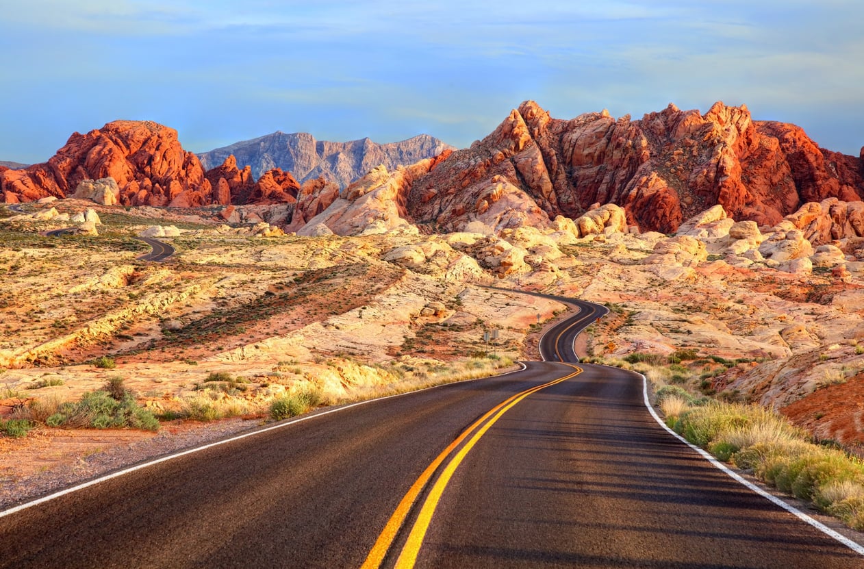 A winding desert road with yellow lines leads toward rugged red and orange rock formations under a partly cloudy sky. Shrubs dot the sandy landscape on both sides of the road.