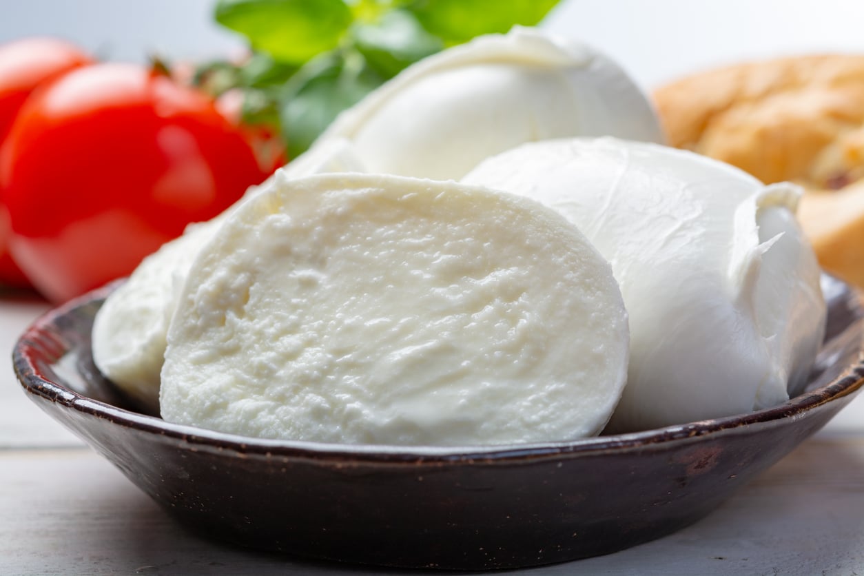 A close-up of fresh mozzarella cheese balls in a dark bowl, with a sliced piece in front. In the background, there are tomatoes, basil leaves, and a piece of bread.