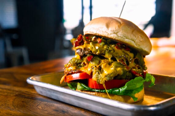 A cheeseburger with melted cheese, lettuce, tomato, and relish on a toasted bun sits on a metal tray on a wooden table.