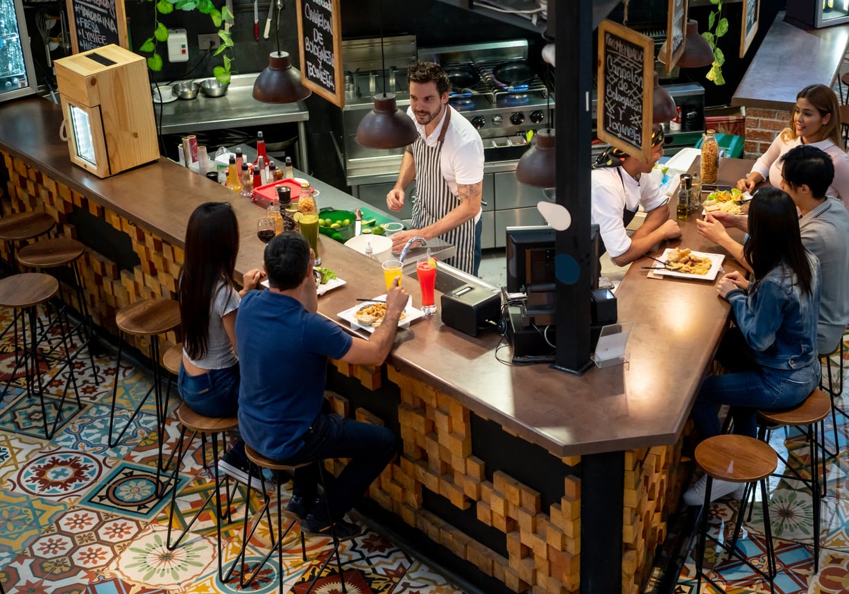 A chef serves food to customers sitting at a curved counter in a lively restaurant with colorful tile flooring and wooden decor.