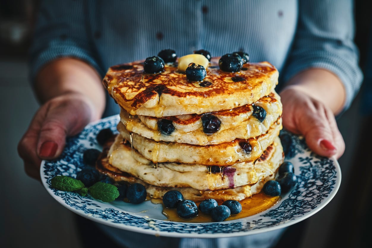 A person holds a plate of stacked pancakes topped with blueberries, butter, and syrup. More blueberries and a mint garnish are on the plate. The person wears a blue striped shirt.