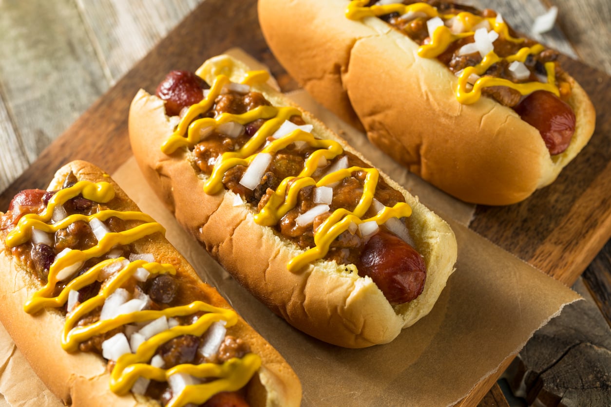 Three chili cheese hot dogs, a shining example of restaurant specialties, are topped with chopped onions and yellow mustard and arranged on parchment paper atop a wooden board.
