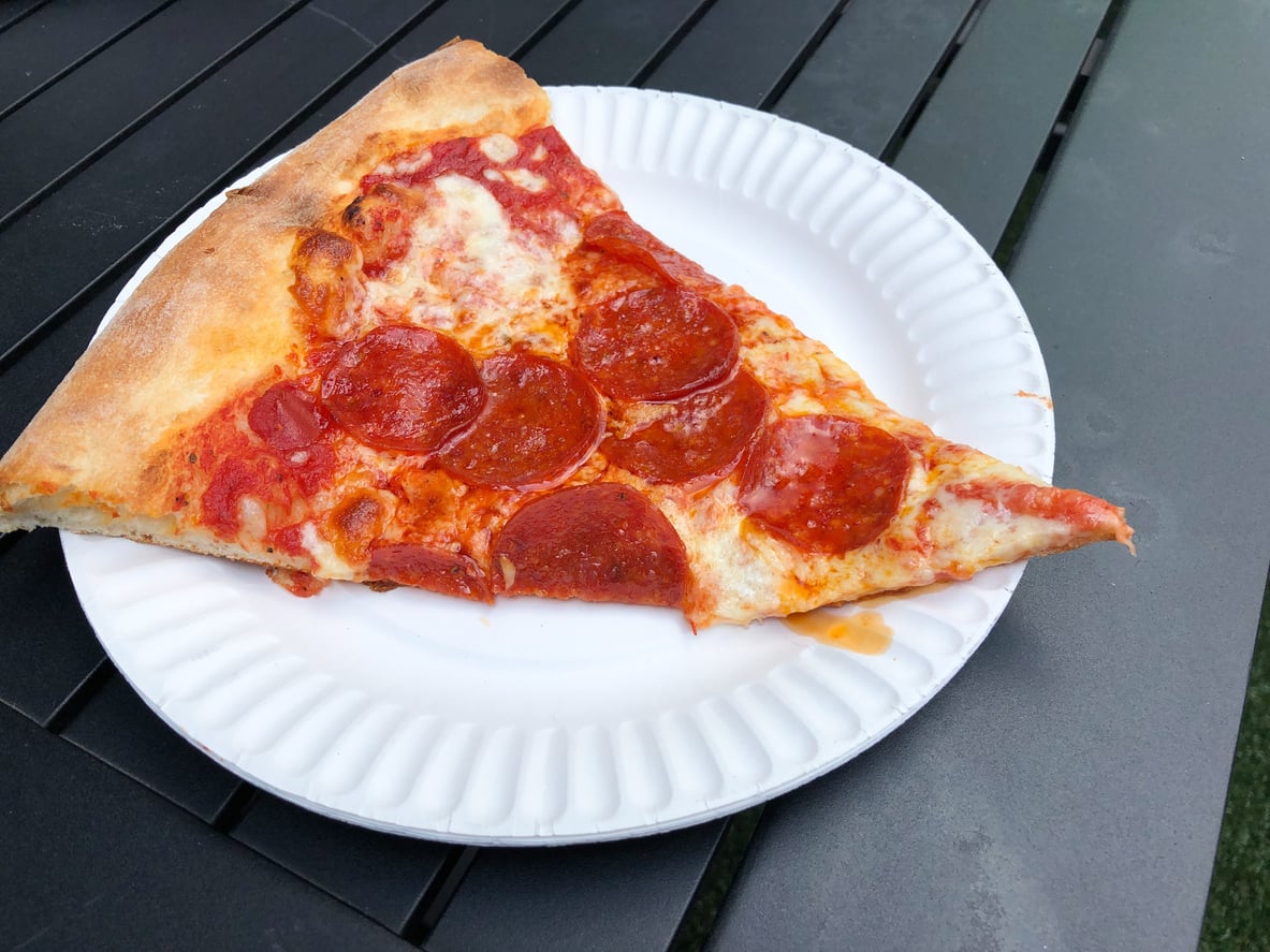 A slice of pepperoni pizza sits on a white paper plate atop a dark outdoor table.