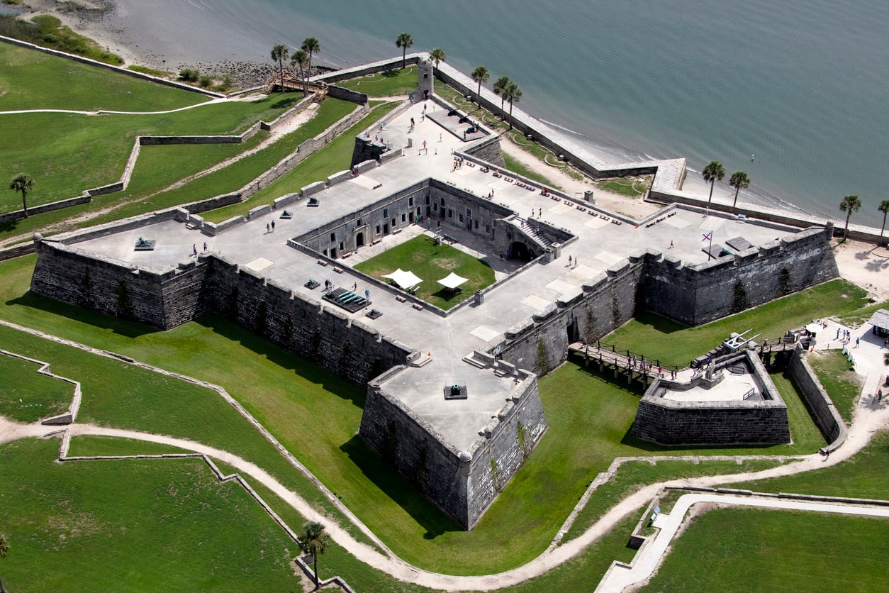 Aerial view of a large, star-shaped stone fortress reminiscent of historic castles, with a central courtyard, surrounded by green grass and water in the background.