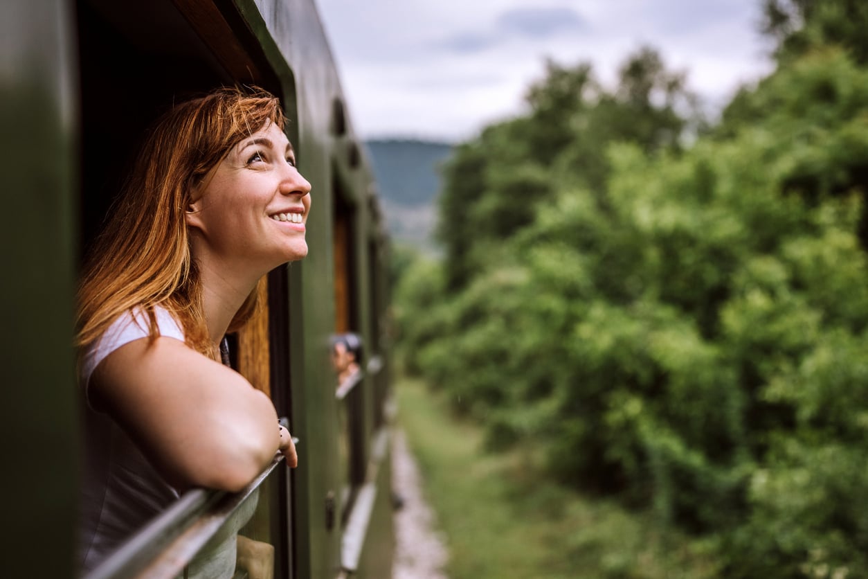A woman leans out of a train window, smiling and looking at the green scenery and trees outside. The sky is cloudy and the train is moving through a forested area.