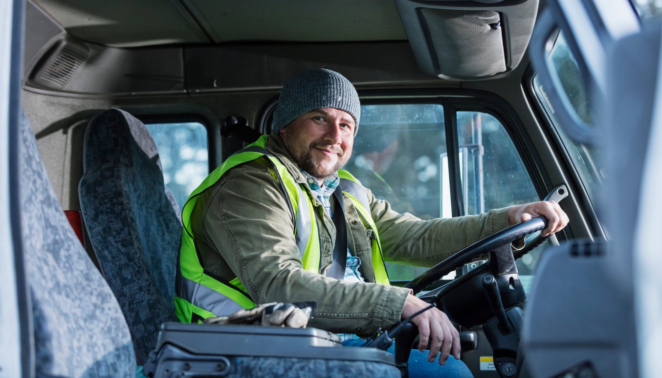 A smiling man wearing a gray beanie and safety vest sits in the driver’s seat of a truck, holding the steering wheel and looking at the camera.