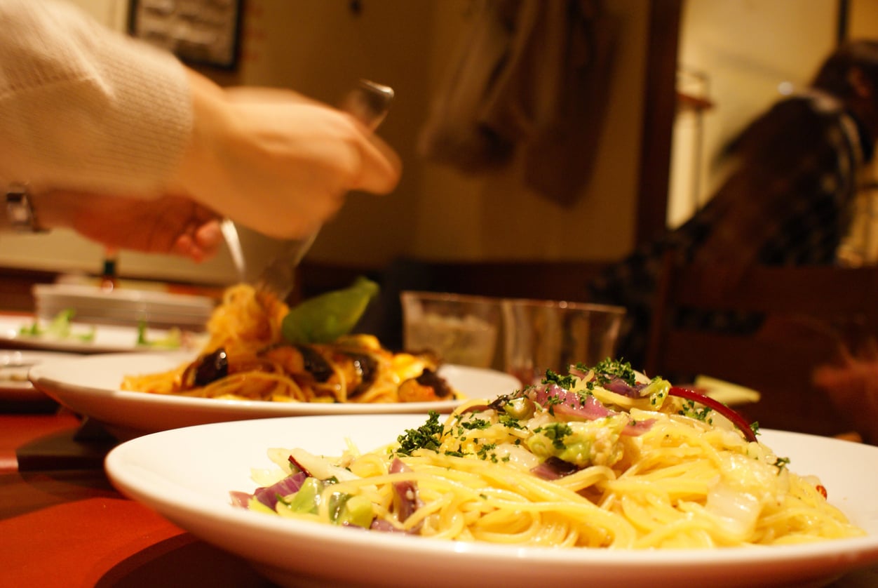 Close-up of two plates of pasta on a red table, one being twirled with a fork by a hand. The pasta is garnished with herbs and vegetables; blurred background of people and restaurant setting.