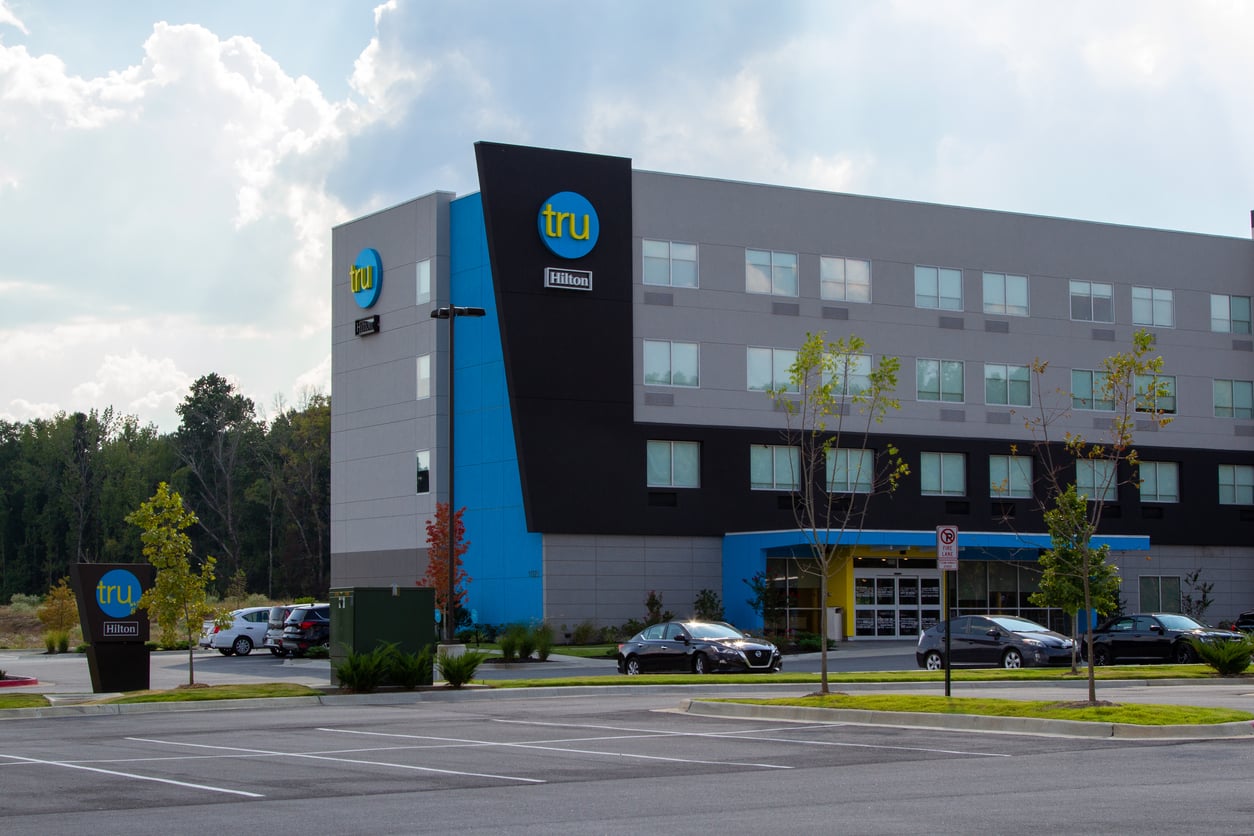 A modern Tru by Hilton hotel building, part of one of the leading hotel chains, features a blue and gray exterior, several cars parked in front, and trees in the background on a partly cloudy day.