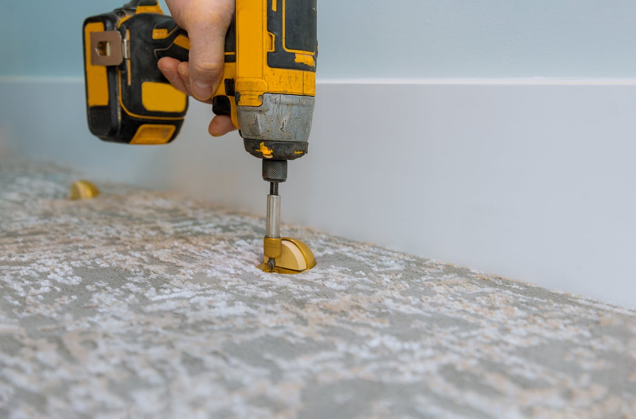 A person uses a yellow and black power drill to secure a floor vent cover onto a carpeted floor next to a white baseboard.