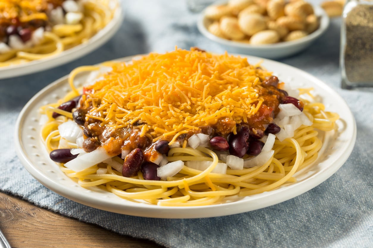 A plate of spaghetti topped with chili, red kidney beans, chopped onions, and shredded cheddar cheese. Bowls of nuts and a pepper shaker are in the background.