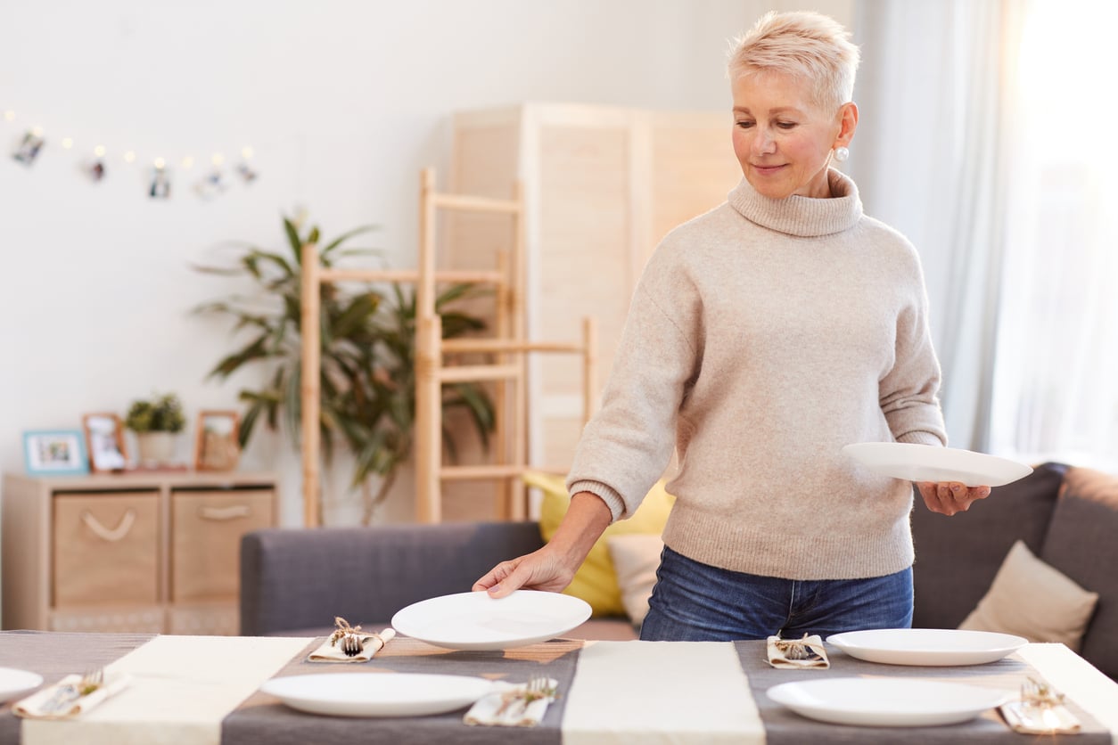 A woman with short blonde hair sets plates on a dining table in a cozy, well-lit living room with neutral decor and plants in the background.