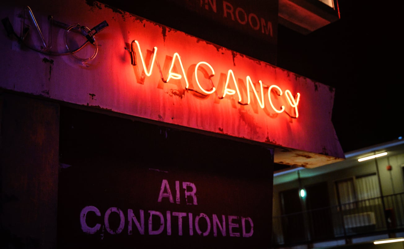 A glowing red neon “VACANCY” sign is lit above a faded “AIR CONDITIONED” sign at night, indicating available motel rooms.