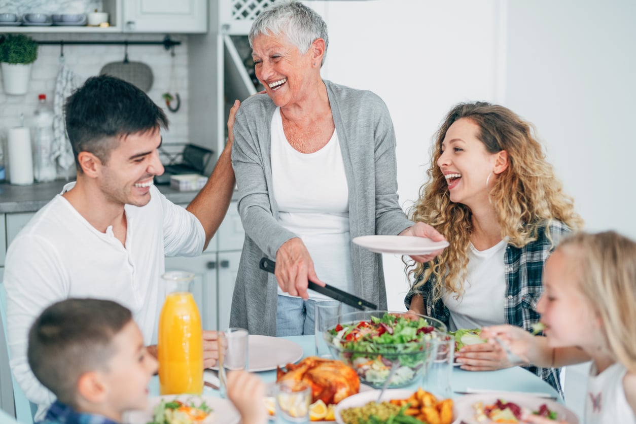 A smiling older woman serving food to a family gathered around a table, laughing and enjoying a meal together in a bright kitchen setting.