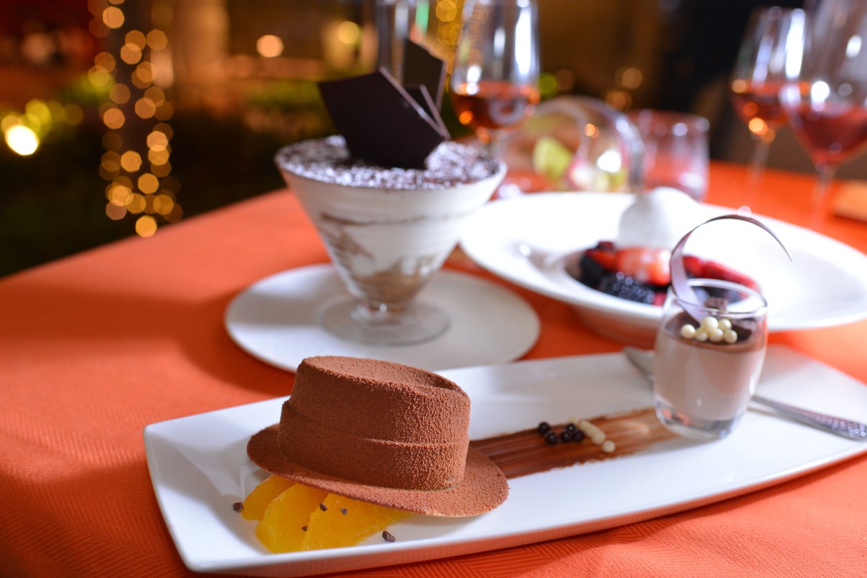 A chocolate mousse shaped like a hat with orange slices, a glass of creamy dessert, and a tiramisu in a martini glass on an orange tablecloth, set for a fancy dinner.