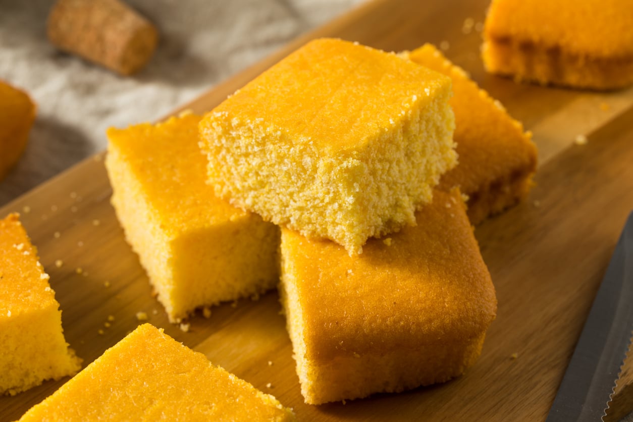 Squares of golden cornbread stacked on a wooden board, showing a moist, crumbly texture. A cork and a cloth are blurred in the background.