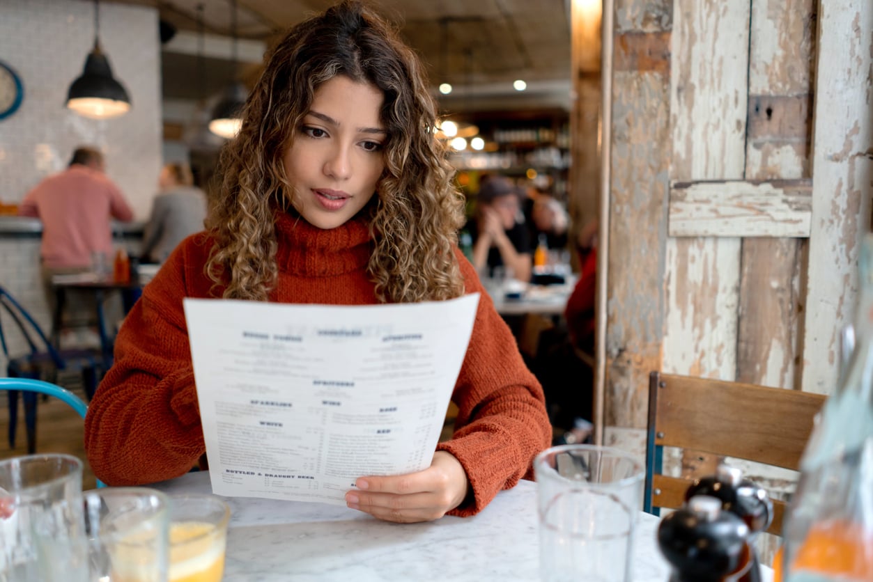 A young woman with curly hair and a rust-colored sweater sits at a table in a cozy café, reading a menu. Glasses of juice and water are on the table. The background shows other people dining.